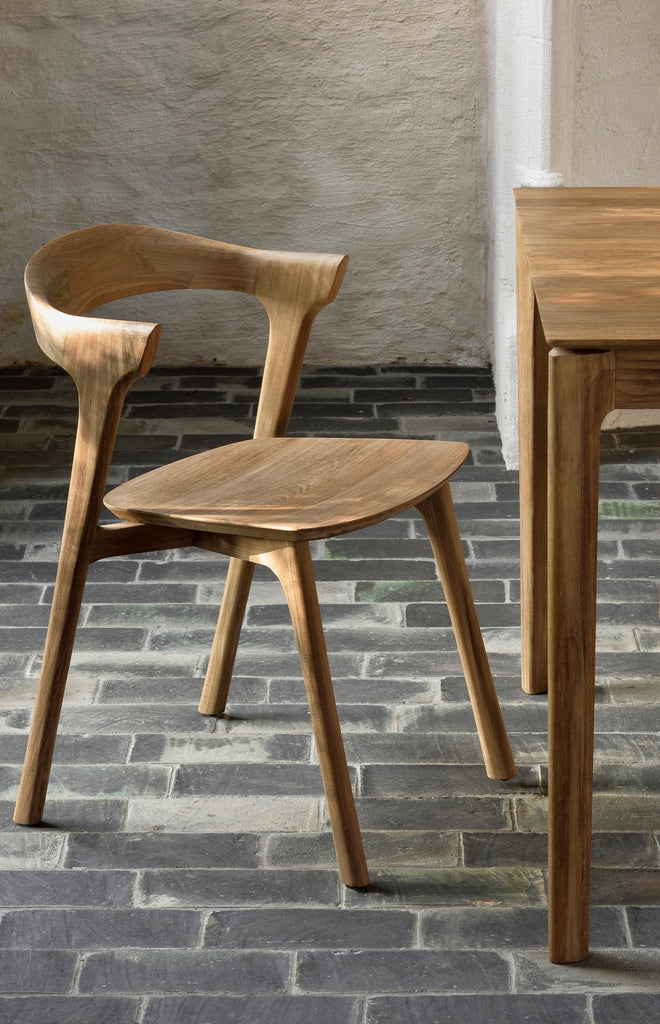 Close up of Teak Bok Dining Chair against charcoal brick floor and plaster walls. - Saffron + Poe, Ethnicraft
