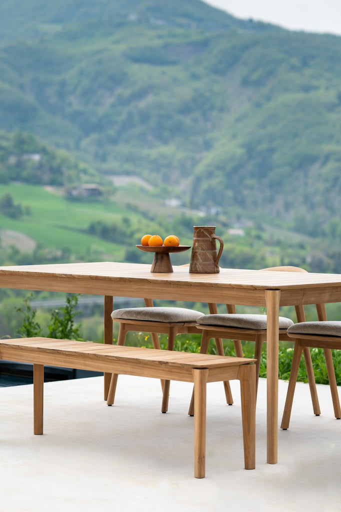 Slatted Teak Bok Outdoor Bench with Teak Bok Outdoor Dining Table on an outdoor patio overlooking a lush, green mountain, styled with a small ceramic bowl and vessel. - Saffron and Poe, Ethnicraft