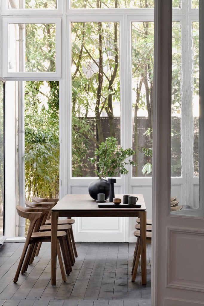 Darkened Teak Bok Dining Table and Darkened Teak Bok chairs in dining room with wall of high windows behind and vase on table with black wood flooring. - Saffron and Poe, Ethnicraft