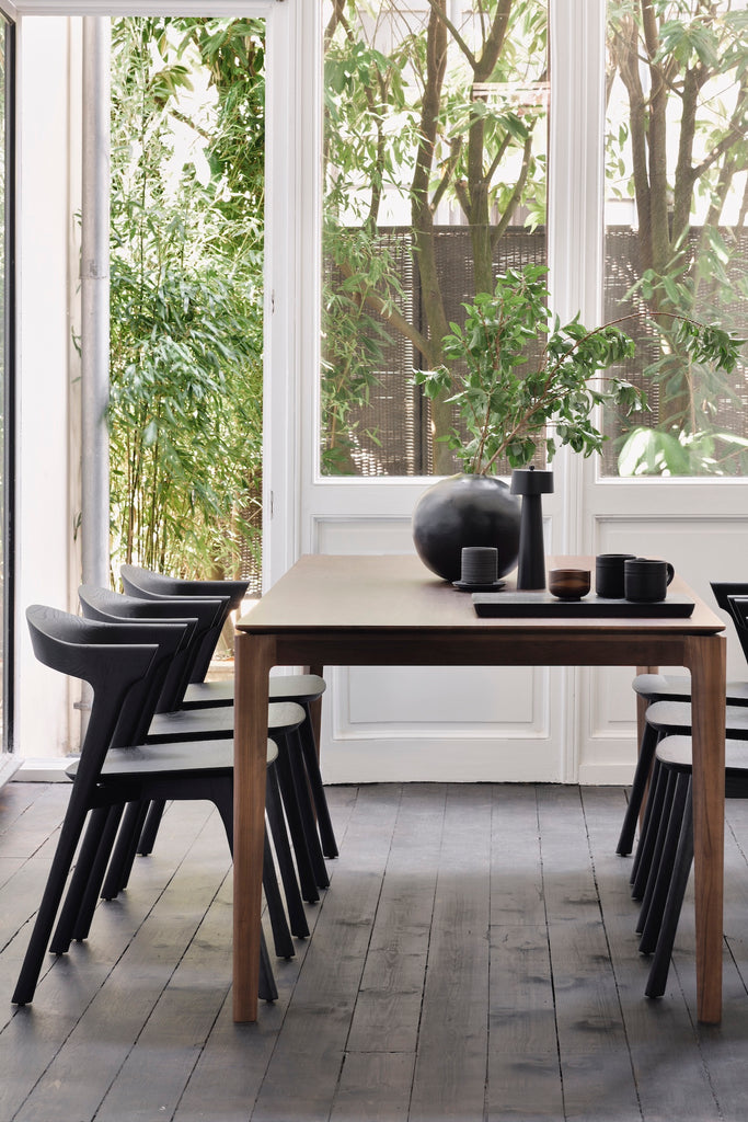 End view of Darkened Teak Bok Dining Table and Black Oak Bok chairs in dining room with wall of windows behind and decorative objects on table and black wood flooring, with floating table top and beautiful wood joinery. - Saffron and Poe, Ethnicraft