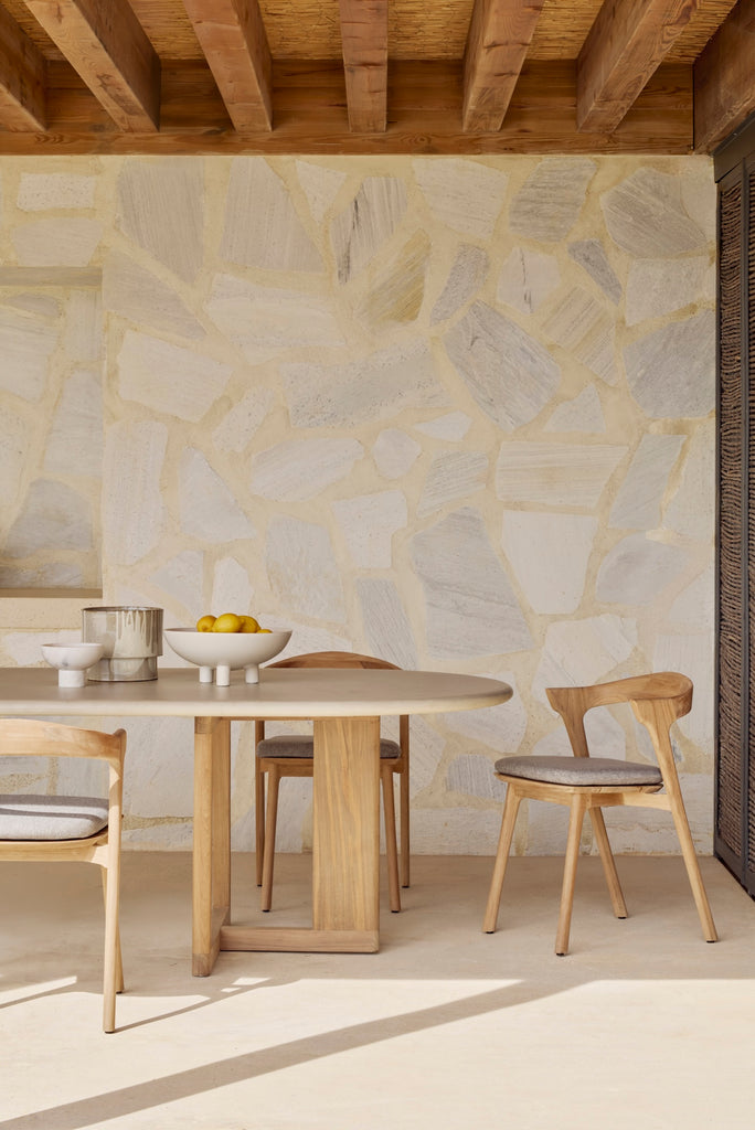 Bok Outdoor Dining Chairs and mocha cushion seat with Trestle Outdoor Dining Table on an outdoor patio with a natural rock wall. - Saffron and Poe, Ethnicraft