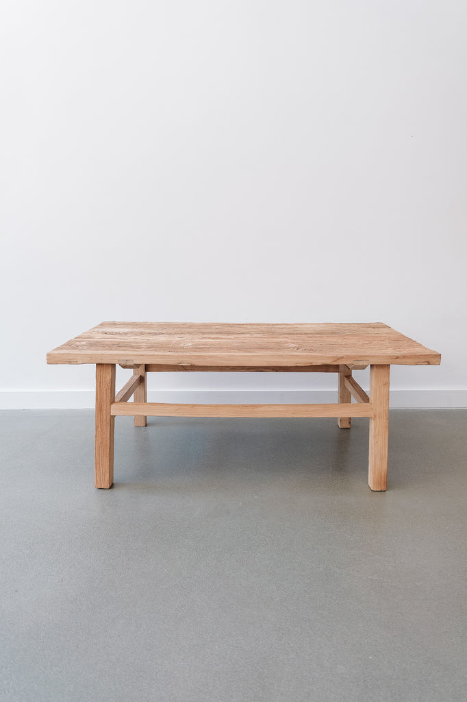 Font view of the Vintage Elmwood Coffee Table in room with white wall and concrete flooring. Crafted with reclaimed light elmwood in a traditional antique Chinese style. - Saffron and Poe.
