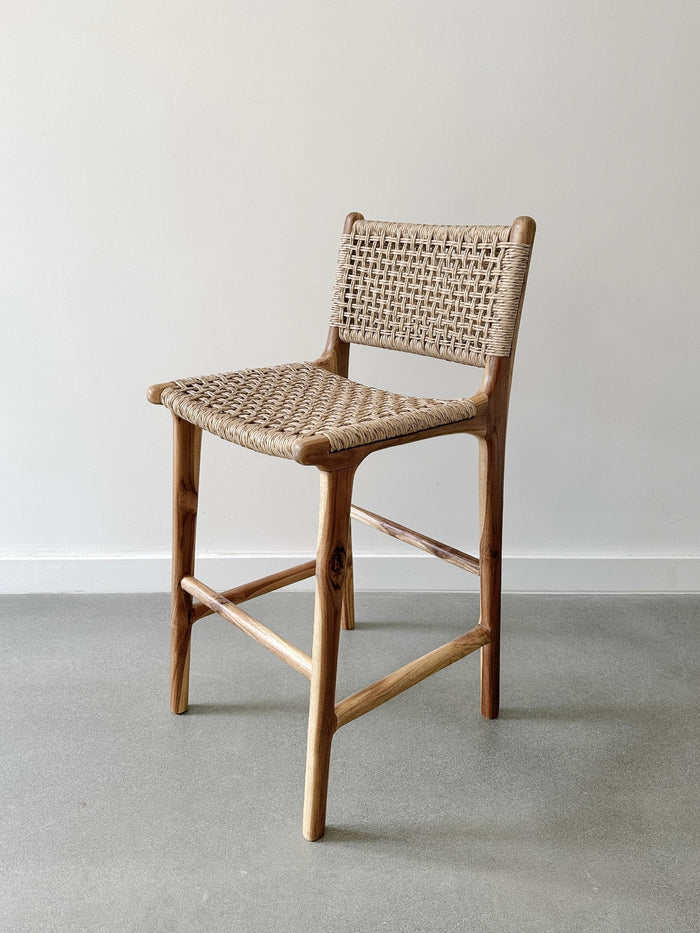 Side product view of the Faux Rattan Outdoor Counter Stool on concrete flooring and a white wall backdrop. - Saffron and Poe