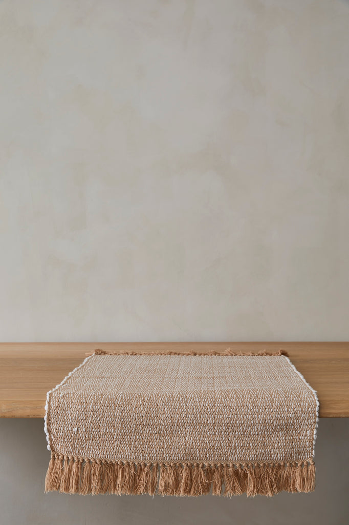 Handwoven bath mat with fringes on a wooden surface against a plain wall.