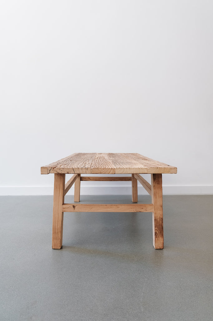 Side view of the Vintage Elmwood Coffee Table in room with white wall and concrete flooring. Crafted with reclaimed elmwood in a traditional antique Chinese style. - Saffron and Poe.