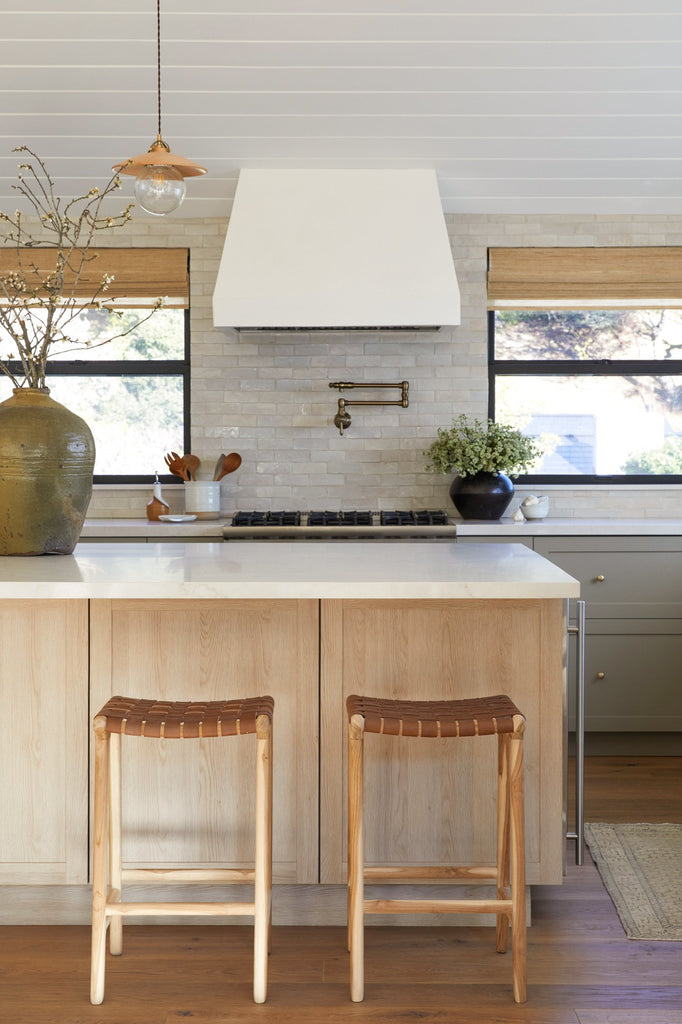 Backless Woven Leather Counter Stool in Saddle styled in a kitchen setting with Antique Clay Pot, Vintage Mijiu Jar, Ceramic Utensil Jar, and Ceramic Olive Oil Bottle. -Saffron and Poe