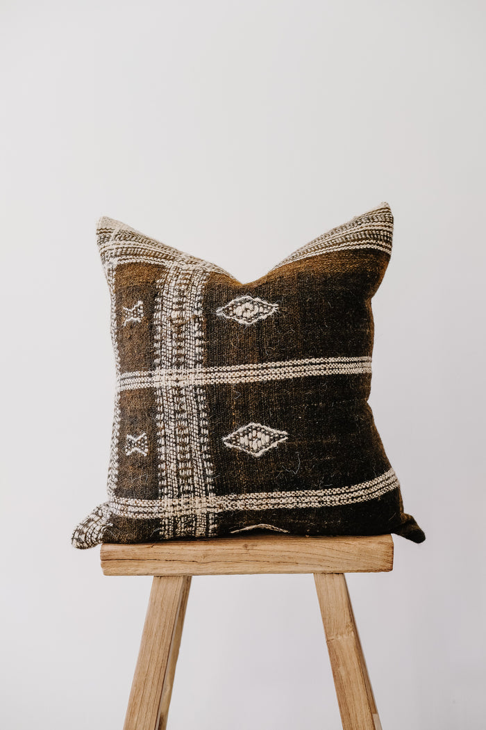 Front view of No. 33 - Handwoven Bhujodi Pillow - Dark Brown on an Antique Stool against a white wall. - Saffron and Poe