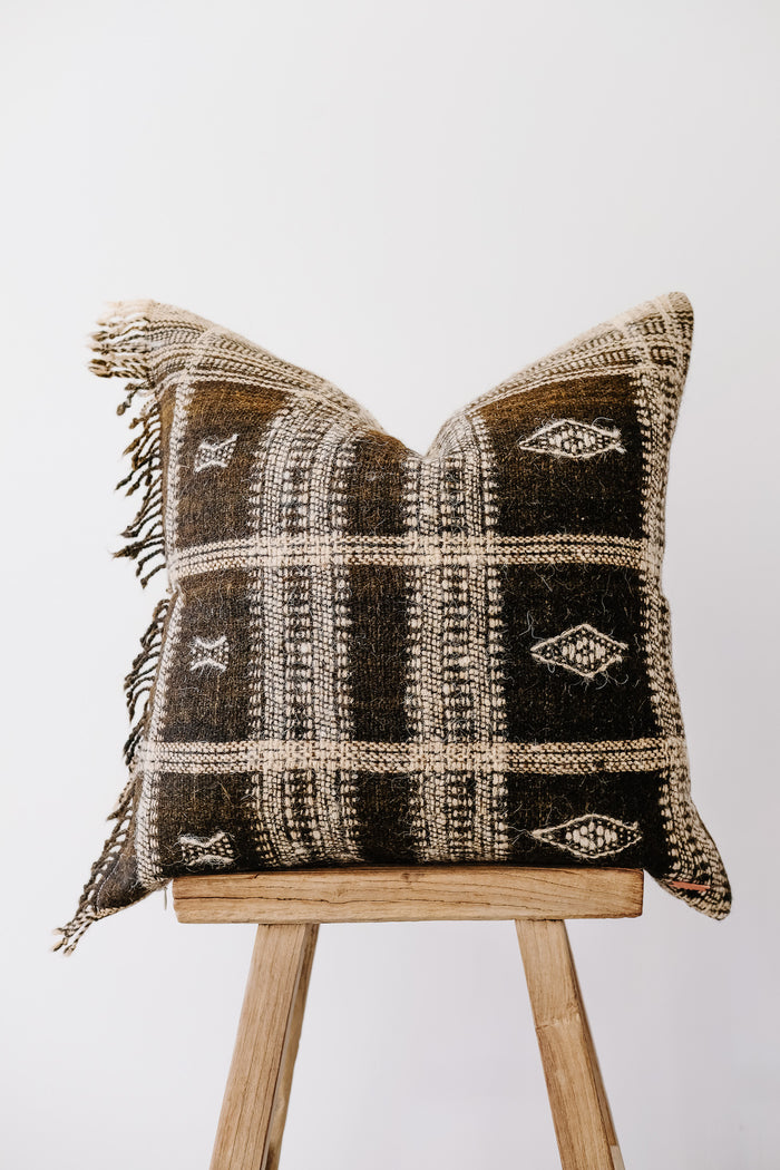 Front view of No. 34 - Handwoven Bhujodi Pillow with Fringe - Dark Brown on an Antique Stool against a white wall. - Saffron and Poe