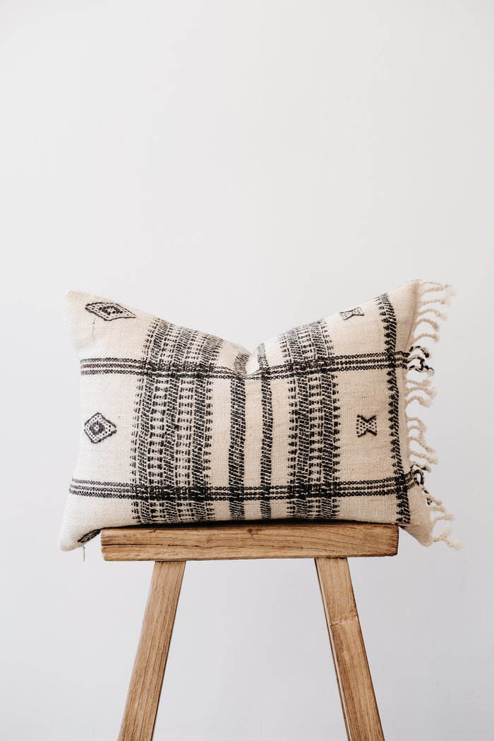 Front view of No. 36 - Handwoven Bhujodi Lumbar Pillow with Fringe - Natural on an Antique Stool against a white wall. - Saffron and Poe