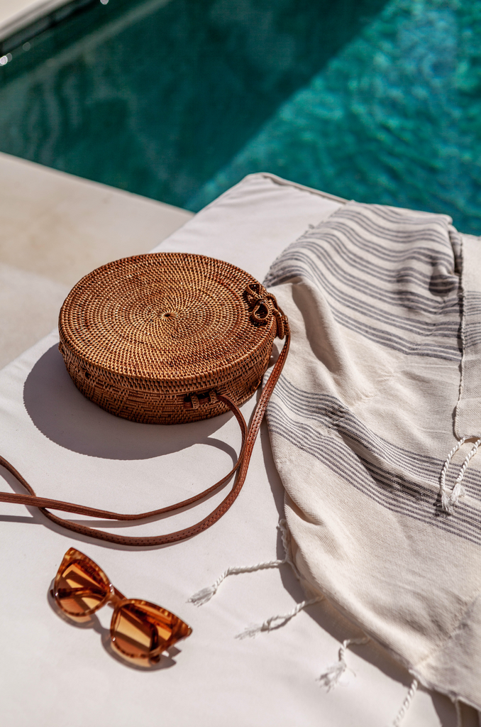 Pool laydown shot with glasses and towel plus casual, chic Tenganan Basket Crossbody Bag Round. Handmade in Tenganan, Bali using natural Ata reed and leather.  - Saffron and Poe