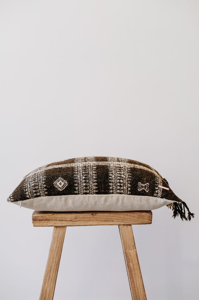 Side view of No. 38 - Handwoven Bhujodi Lumbar Pillow with Fringe - Dark Brown on an Antique Stool against a white wall. - Saffron and Poe