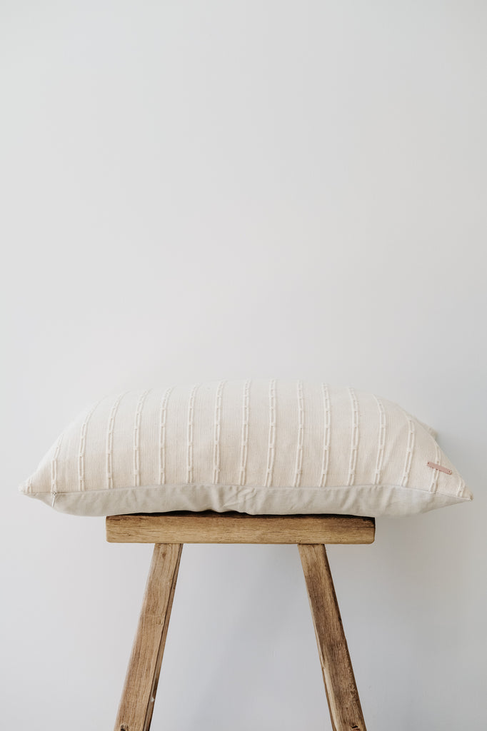 Side view of No. 05 - Ivory with Link Pattern Embroidered Lumbar Pillow on a Chinese Stool against a white wall. - Saffron and Poe