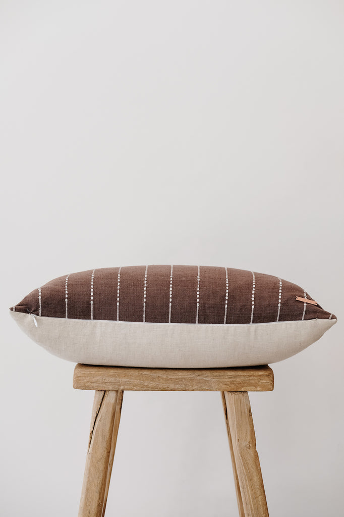 Side view of No. 14 - Hmong Hemp Batik Pillow - Dark Brown on an Antique Stool against a white wall. - Saffron and Poe