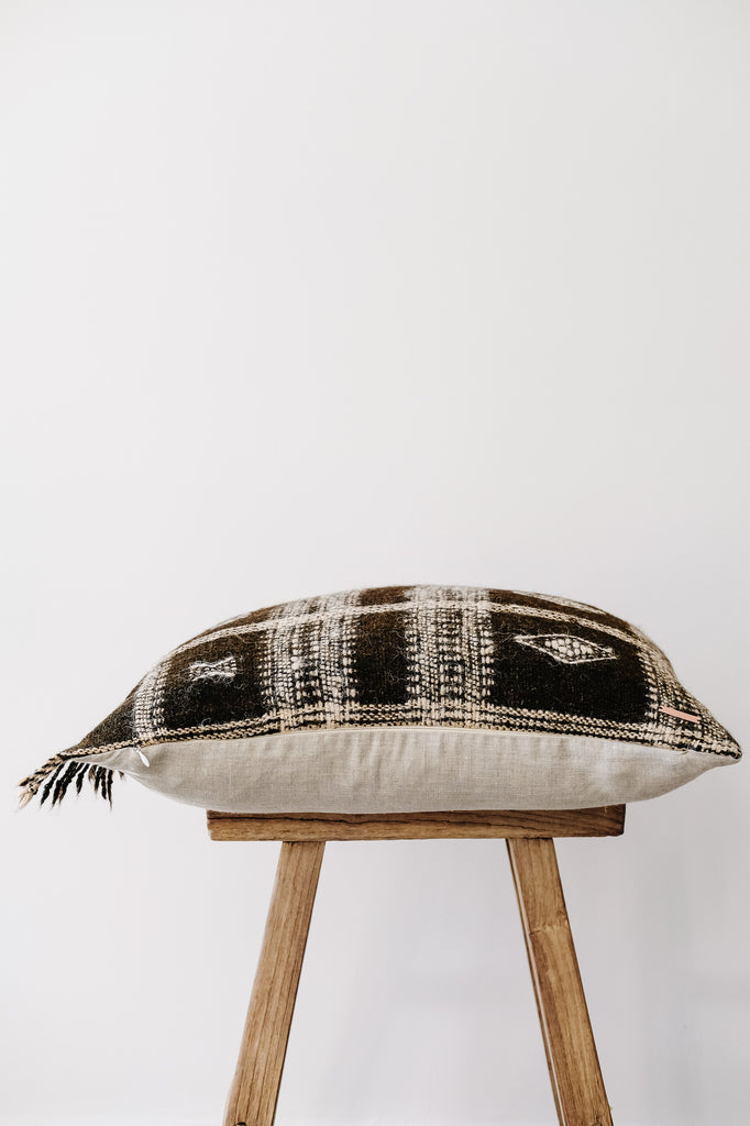 Side view of No. 34 - Handwoven Bhujodi Pillow with Fringe - Dark Brown on an Antique Stool against a white wall. - Saffron and Poe
