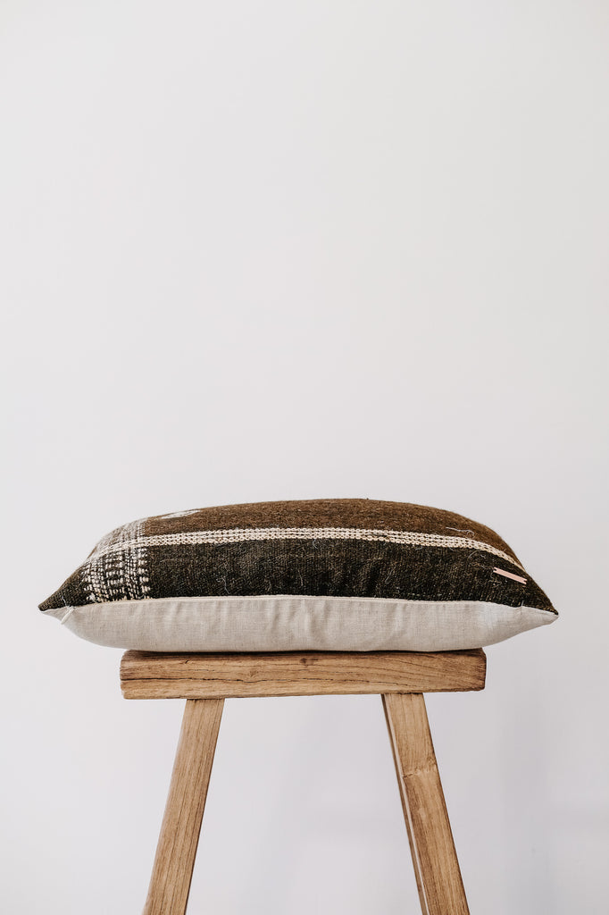 Side view of No. 39 - Handwoven Bhujodi Lumbar Pillow - Dark Brown on an Antique Stool against a white wall. - Saffron and Poe