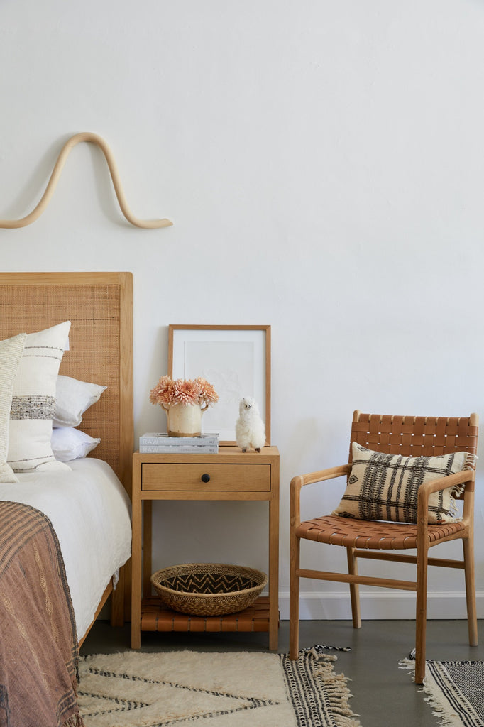 Styled angled view of the Woven Leather Strap Dining Arm Chair in beige next to the Oak Side Table and Rattan headboard. - Saffron and Poe.