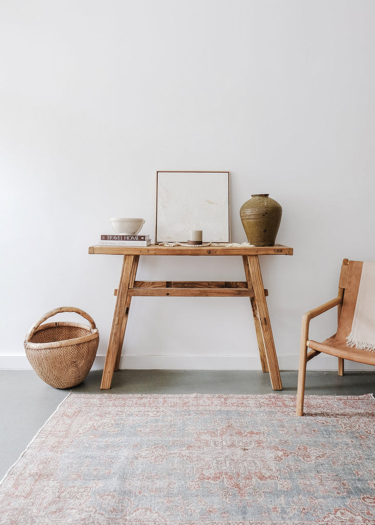 Styled Terrazzo Bowl on an Antique Console Table with a Leather Sling Lounge Chair, Bali Beads, and an Antique Chinese Basket on a Vintage Turkish Oushak Rug against a white wall. - Saffron and Poe