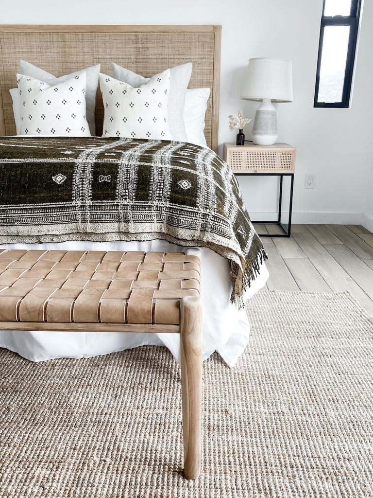 Front half shot of styled bed with hemp textile pillows, nightstands, hardwood floors, vase, lamp, woven leather strap bench in beige, jute area rug, and dark brown Indian bhujodi bed throw on bed.