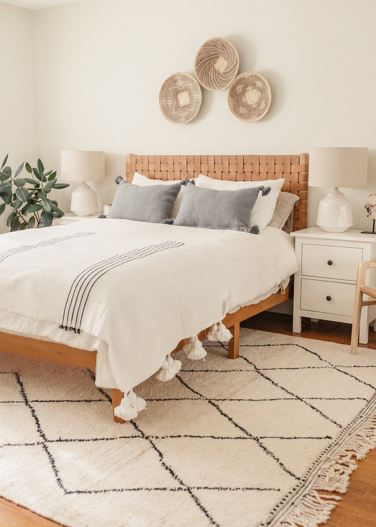 Styled Grey Moroccan Pom Pillow with a Woven Leather Strap Headboard and Ilala Palm Wall Baskets and a Leather Sling Lounge Chair in a bright bedroom. - Saffron and Poe