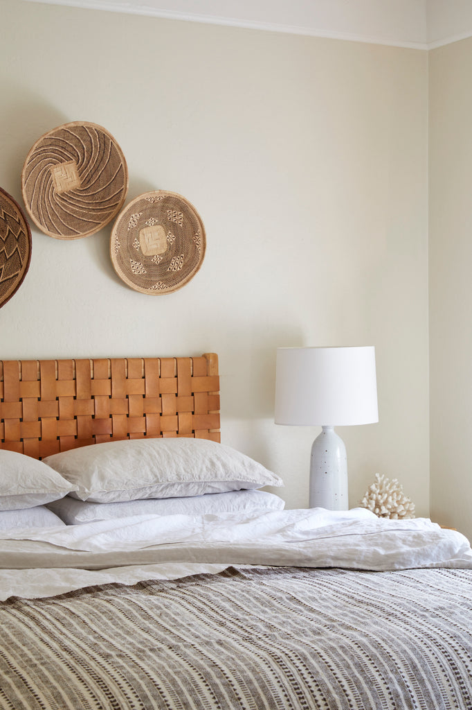 Styled view of White Linen Sheet Set with a Handwoven Bhujodi Blanket - Natural, a Woven Leather Strap Headboard, a Hand Thrown Ceramic Lamp, and Ilala Palm Wall Baskets. - Saffron and Poe