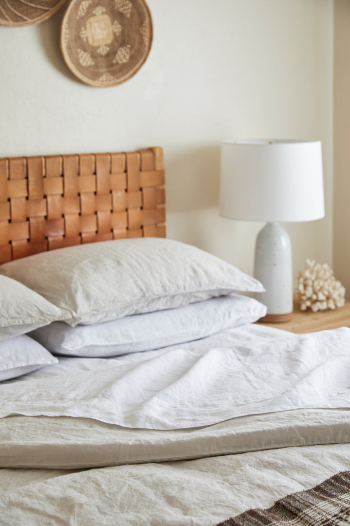Styled view of White Linen Sheet Set with a Handwoven Bhujodi Blanket - Natural, a Woven Leather Strap Headboard, a Hand Thrown Ceramic Lamp, and Ilala Palm Wall Baskets. - Saffron and Poe