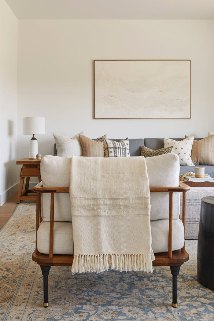 Styled view of our Handwoven Colombian Throw in ivory draped over a decorative chair in a living room setting which includes our Ceramic Lamp, Vintage End Table, Bhujodi pillows, and Kaleen Cameron art work. - Saffron and Poe.