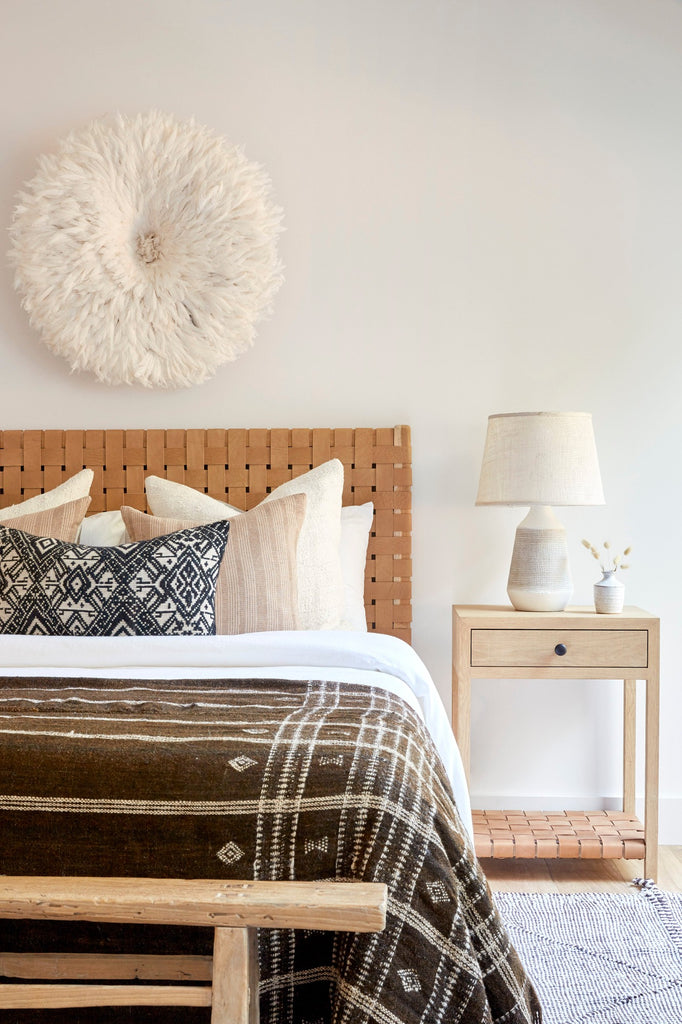 Handwoven Bhujodi Blanket in Dark Brown in a bedroom setting with Woven Leather Headboard, Beige Embroidered Pillows, Antique Miao Lumbar Pillow, Antique Bench, and White Oak Side Table. - Saffron + Poe