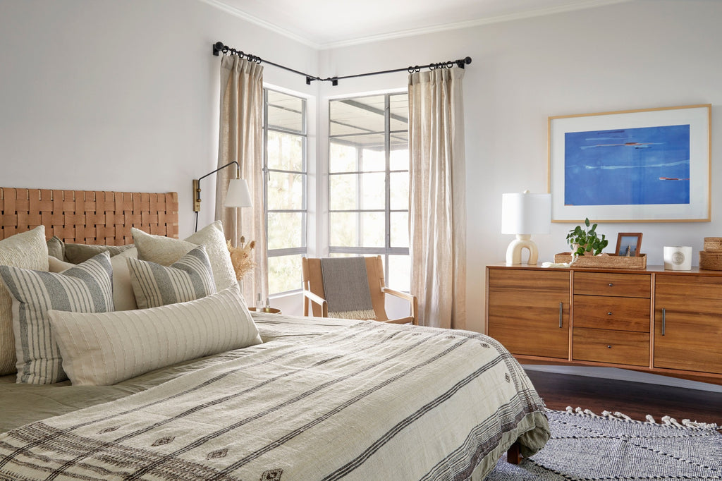 Handwoven Bhujodi Blanket in Natural in a bedroom setting with Woven Leather Headboard, Ivory Handwoven Columbian Pillows, Ivory Embroidered Pillows, Ivory Extra Long Lumbar Pillow, Leather Sling Lounge Chair, and Moroccan Flat Weave Kilim Rug. - Saffron + Poe