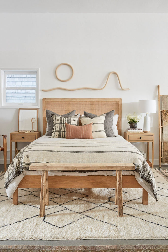 Handwoven Bhujodi Blanket in Natural in a bedroom setting with Rattan Headboard, Antique Bench, White Oak Side Table, Katie Gong Sun and Mountain art piece, Ceramic Lamp, Bhujodi Pillows, and Beni Ourain Rug. - Saffron + Poe