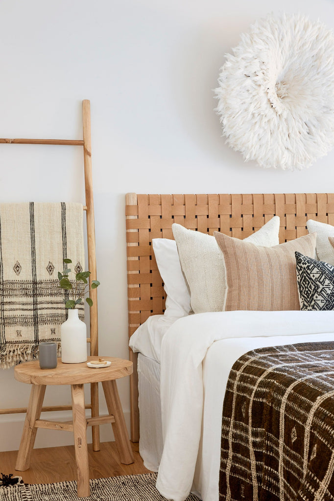 Woven Leather Strap Headboard in a bedroom setting with Vintage Round Side Table, Natural Bhujodi Throw, Dark Brown Bhujodi Blanket, Beige Embroidered Pillow, and. Antique Miao Lumbar Pillow. -Saffron + Poe