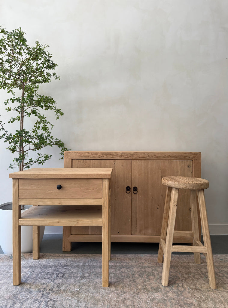 Vintage cypress and elmwood furniture including a side table, cabinet, and stool in a room with a black olive tree. Saffron + Poe