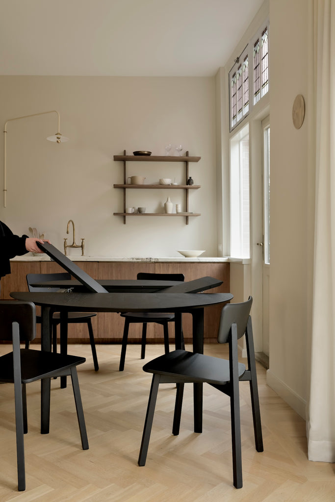 Black Oak Bok Round Extendable Dining Table and Black Oak Bok chairs in kitchen with herringbone wood flooring, with person showing extension leaf mechanism, featuring float top and beautiful wood joinery. - Saffron and Poe, Ethnicraft.