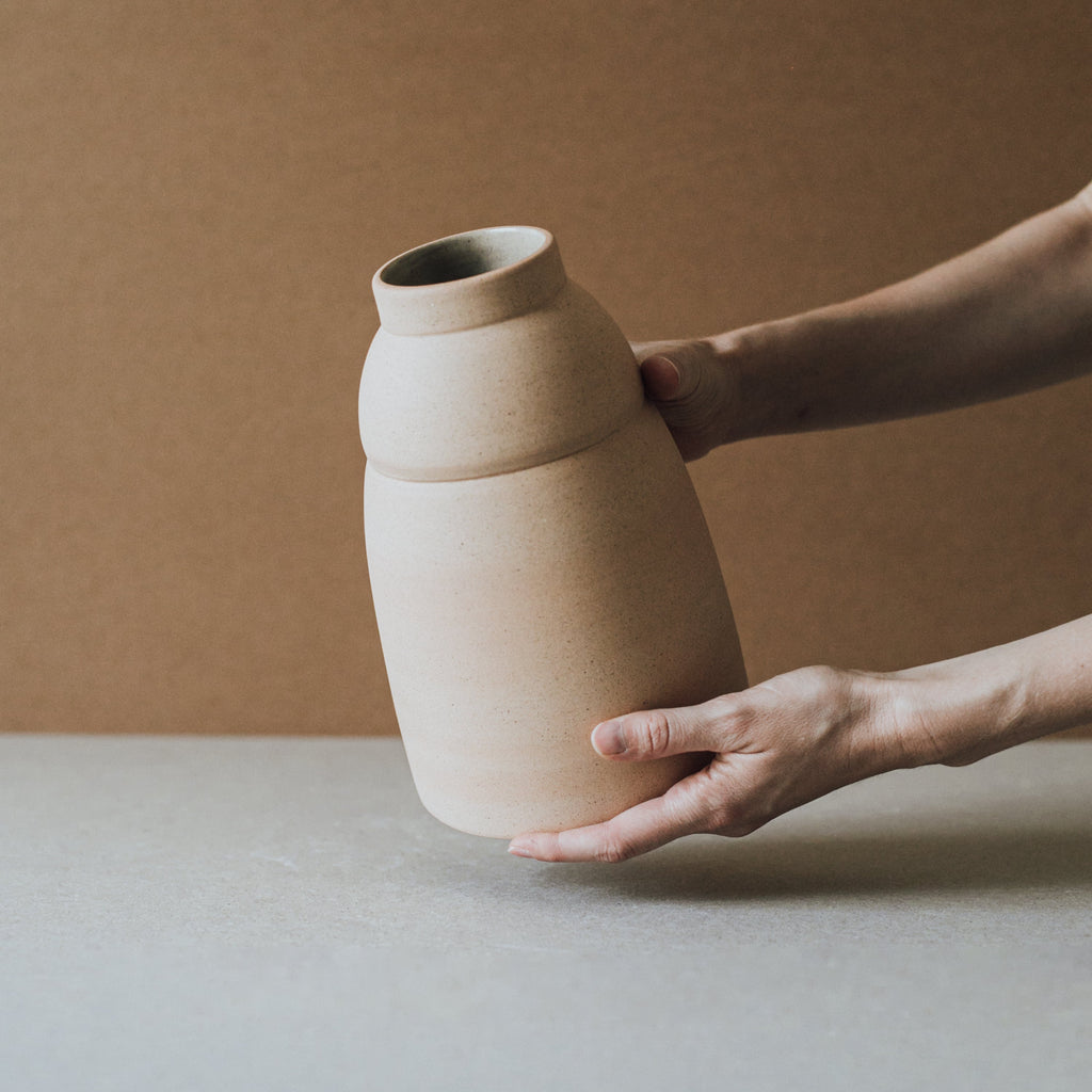 Person holding a beige ceramic vase against a neutral background