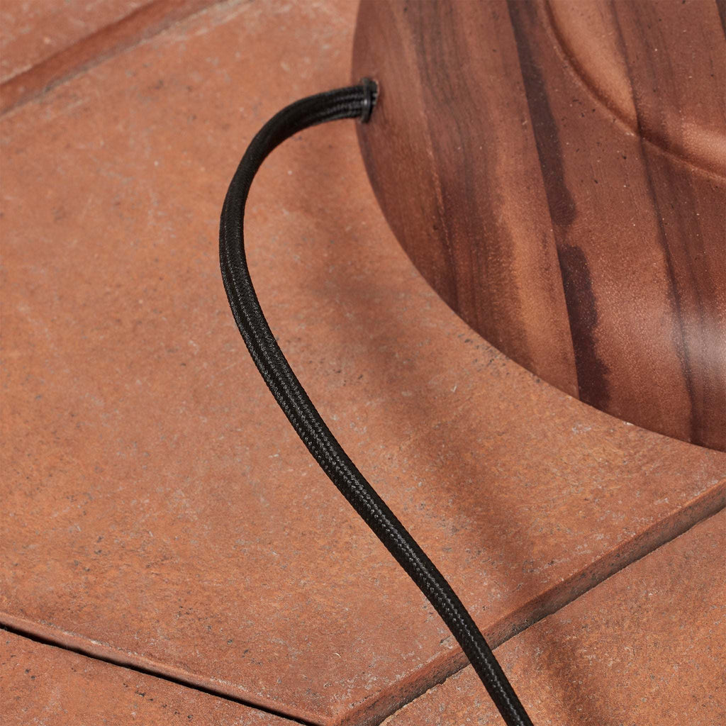 Black cable of the Alhambra Floor Lamp running along a wooden surface with a red-brown stone-like texture.