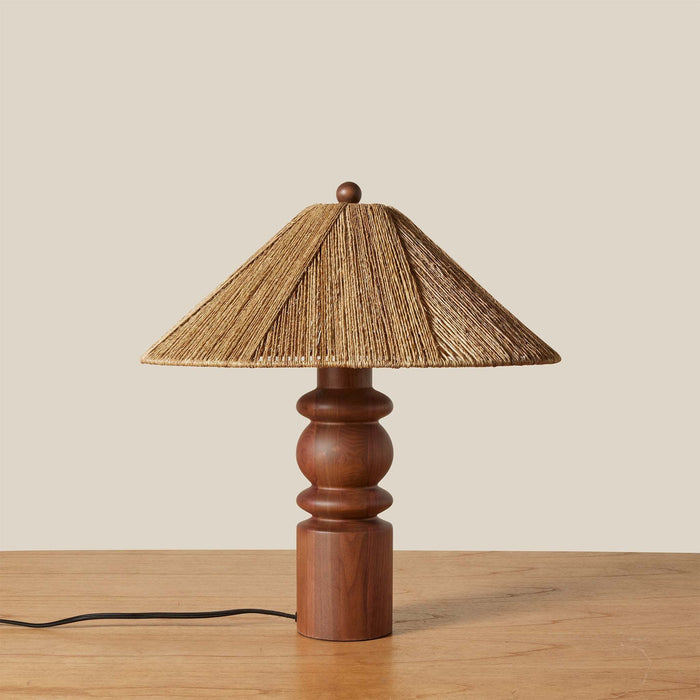 An Alhambra table lamp with a warm brown wooden turned base and a natural beige woven straw shade, set on a light wood tabletop against a solid beige background.