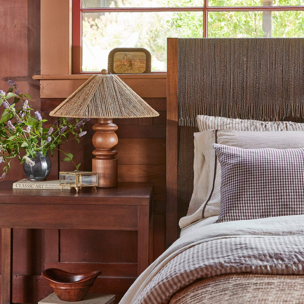 A rustic bedroom scene featuring a wooden nightstand with a woven lampshade Alhambra table lamp, a vase of purple flowers, and a bed with a textured brown headboard, striped bedding, and a purple gingham pillow.