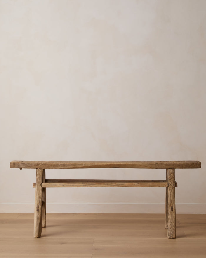 A rustic wooden bench with a thick plank seat and a lower support rail, shown against a plain beige wall and light wood floor.