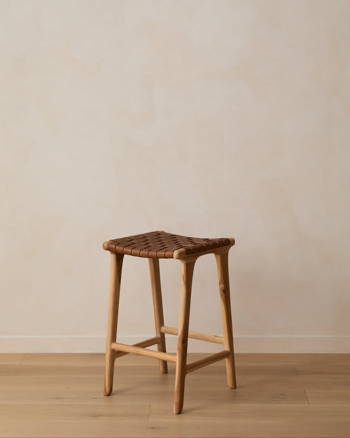Backless woven leather counter stool on oak floor against limewashed wall. Saffron + Poe