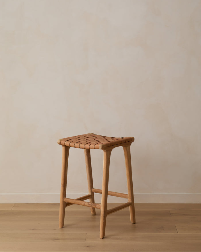 Angle view of Beige Woven Leather Counter Stool on oak wood floor in front of limewashed wall. Saffron + Poe