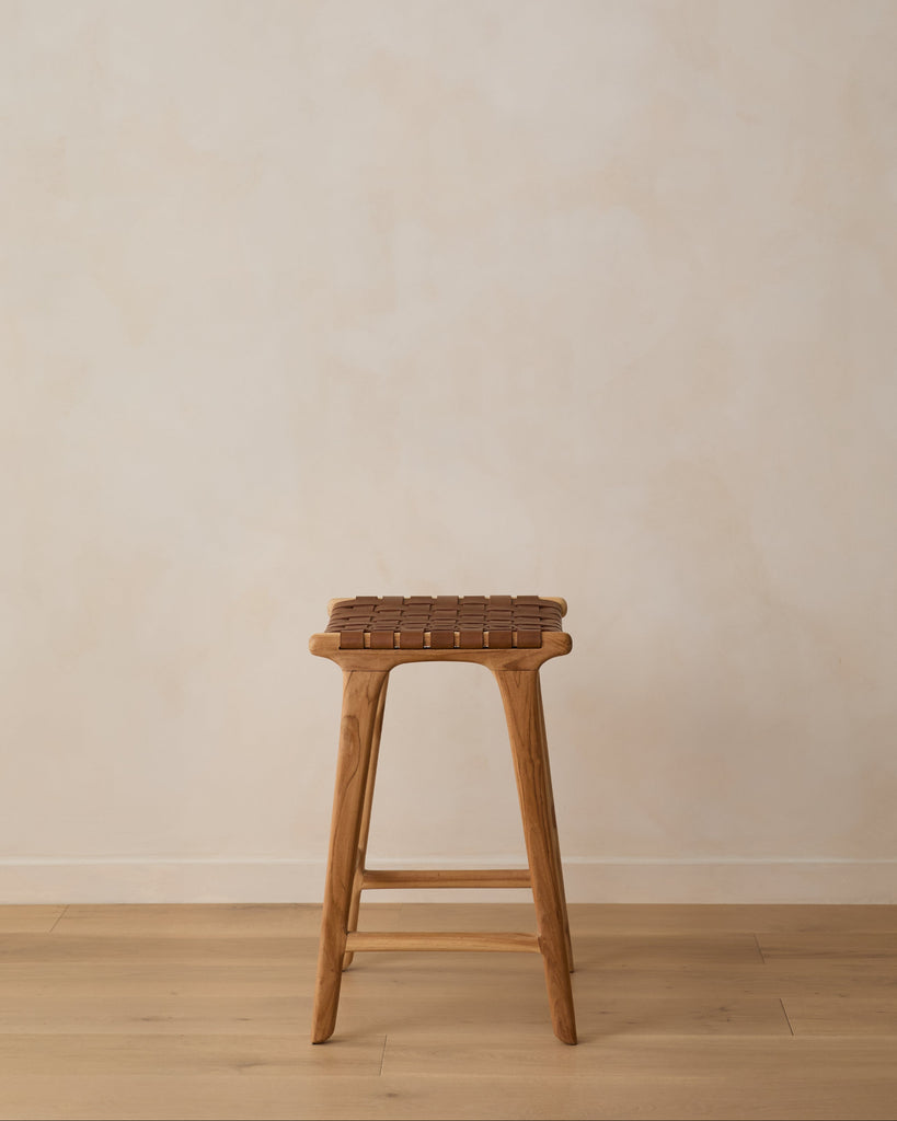 Side angle of backless woven leather counter stool on oak floor against limewashed wall. Saffron + Poe