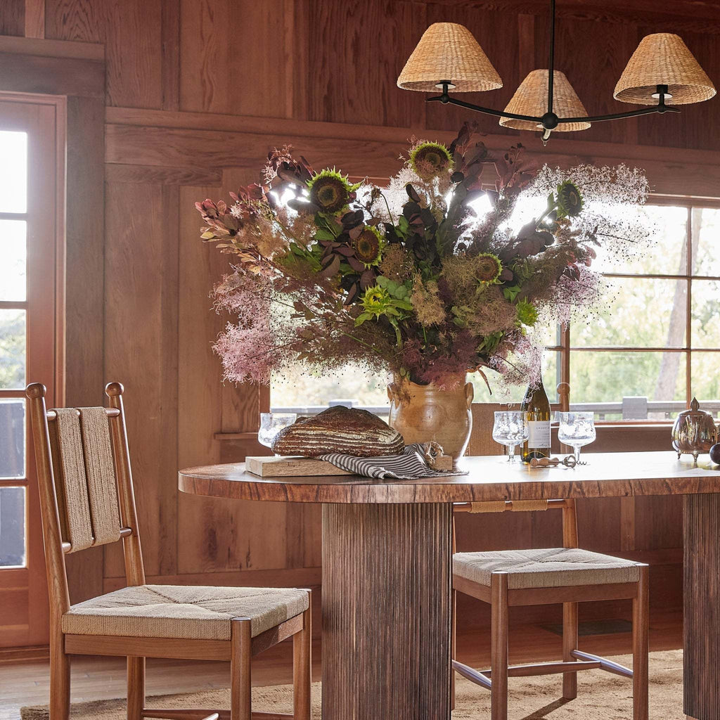 A rustic dining room scene with a large floral arrangement in a vase on a wooden table, with a Bainbridge Chandelier, two wooden chairs and stools, and wood-paneled walls in the background.
