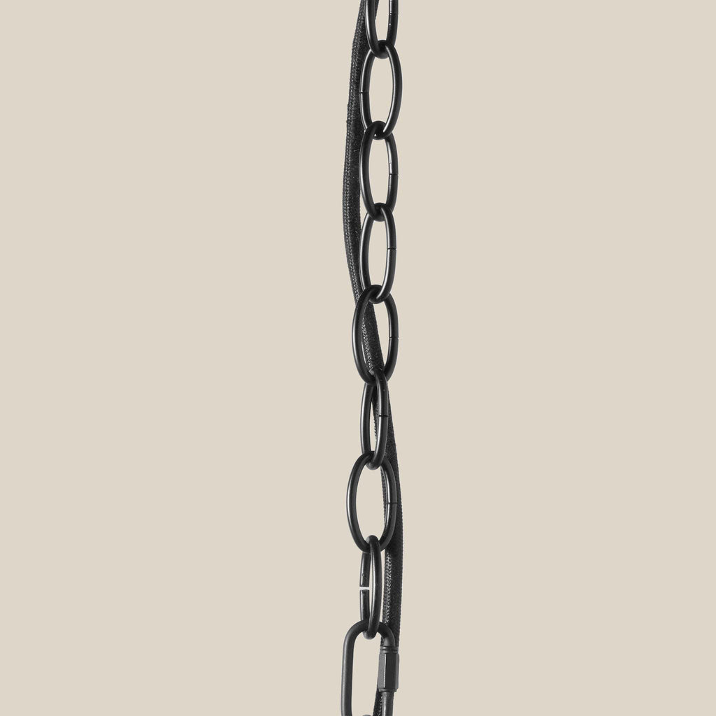 A close-up of a black metal chain with elongated oval links and a carabiner-style connector, shown vertically against a plain warm beige background.