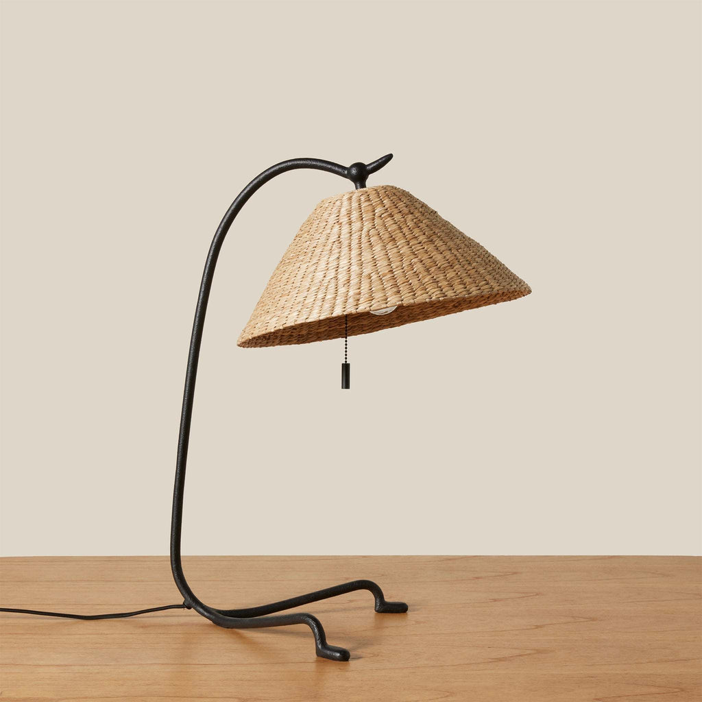 Bainbridge table lamp with wicker shade and black metal base on a wooden surface.