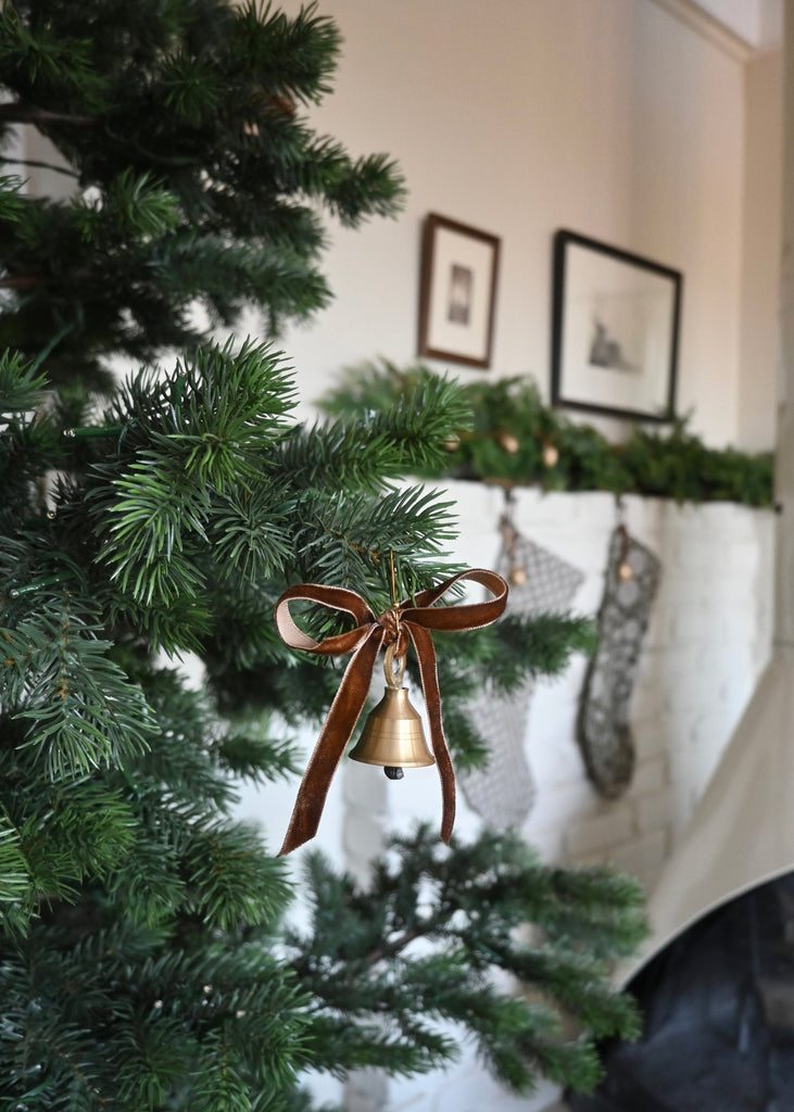 Decorative bell ornament with a brown ribbon on a Christmas tree, with a fireplace and stockings in the background.
