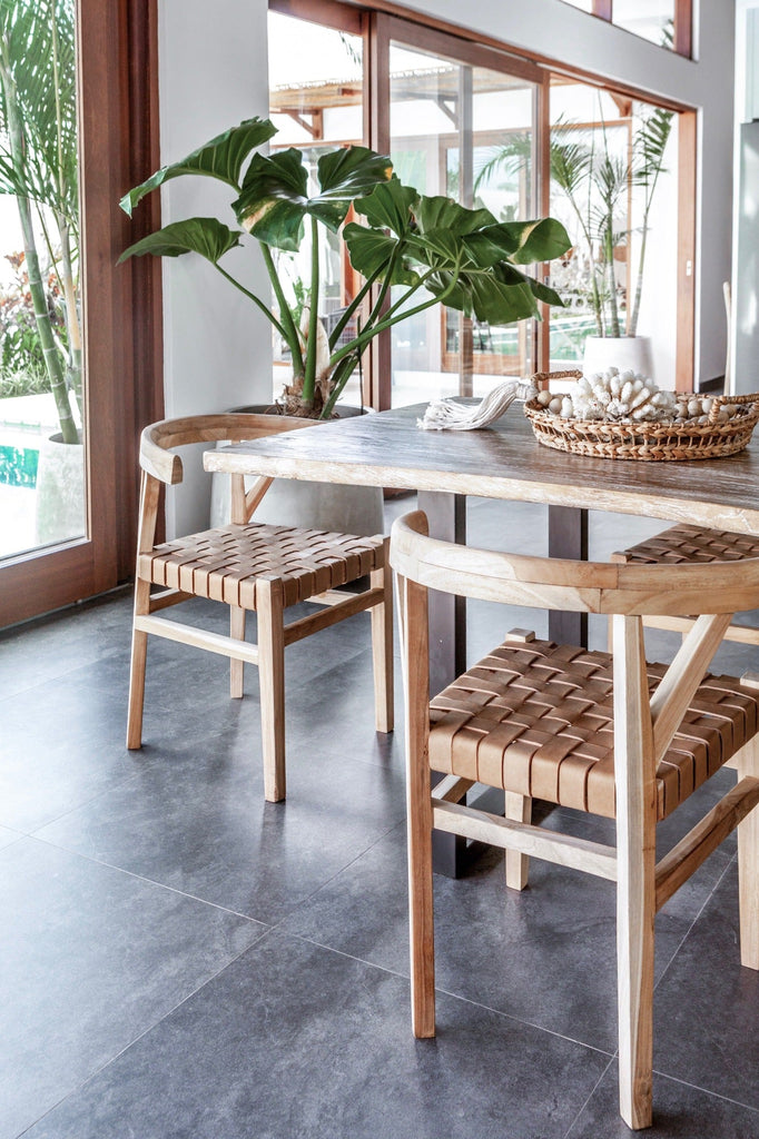 Two curved wood and woven beige leather dining chairs inspired by the wishbone Chair at the kitchen table with a plant. Handcrafted in Bali with Teak wood and vegetable-tanned leather imported from Java. - Saffron and Poe