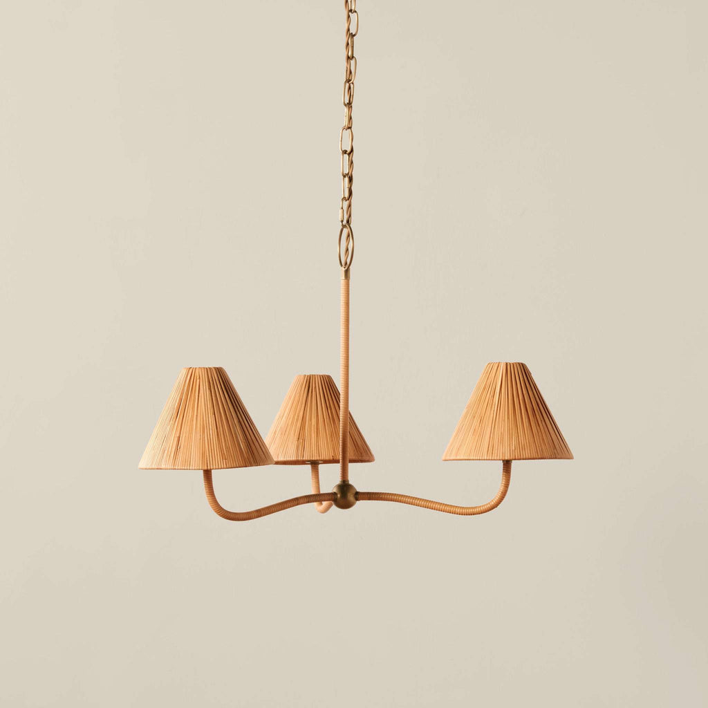 A three-light Delphine chandelier with a curved brass metal frame and pleated beige fabric shades, suspended from a chain against a solid light beige background.