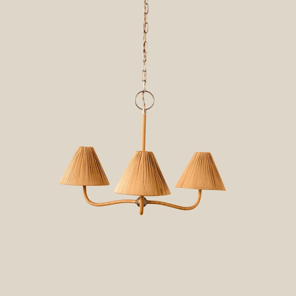 A three-light Delphine chandelier with pleated beige fabric shades and a curved brass-toned metal frame, suspended by a chain against a solid light beige background.