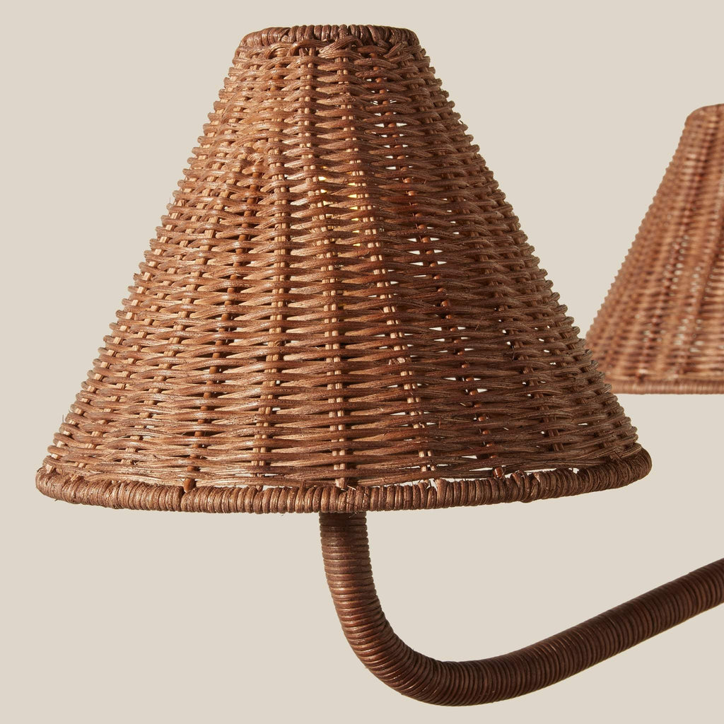 Close-up of woven rattan Delphine chandelier lampshade in a warm medium brown tone, attached to a matching curved arm, with a second lampshade partially visible in the background against a solid light beige backdrop.