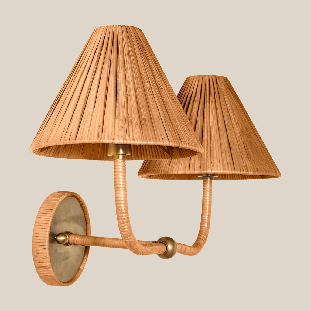 A two-light Delphine wall sconce with pleated beige rattan shades, curved rope-wrapped arms, and a round textured backplate on a solid light beige background.