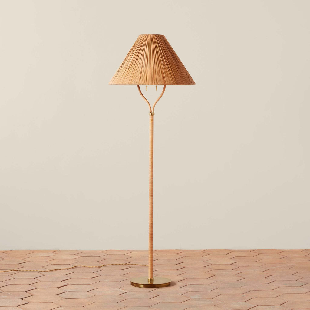 A Delphine floor lamp with a pleated conical shade, a slender bamboo-like stem, and a round brass base, standing on a terracotta tile floor against a plain beige wall.